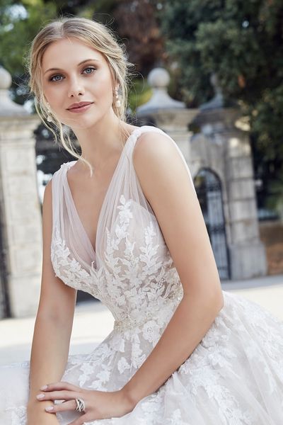 Close up of a model sat by grand black iron gates in Ronald Joyce 18507, a tulle ballgown wedding dress with lace embroidery detail, ruched tulle straps and a matching v-neckline